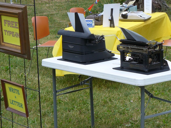 Rear view of table with two old typewriters. To left hang two sings reading Free Type-In and Write a Letter