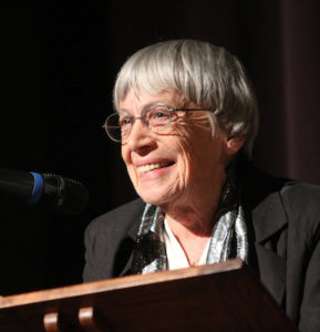 Ursula K. Le Guin. An older Caucasion woman with short white hair, face with creases, wearing glasses, stands at a podium. She is smiling genially.