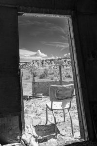 Black and white view out of the door of an old RV trailer. View is a desert with a beat-up metal chair in foreground, wooden fence midground, and sagebrush hills with sky and clouds above in background