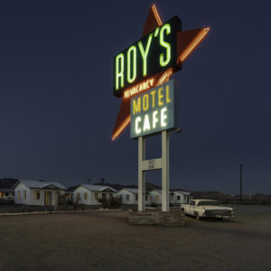 Outdoor shot of old neon hotel sign. Roy's Motel. At base of sign are 3 small house-shapes and an old car.
