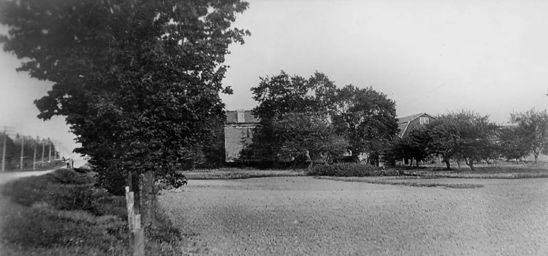 Cream brick farm house and barn seen from a distance looking east