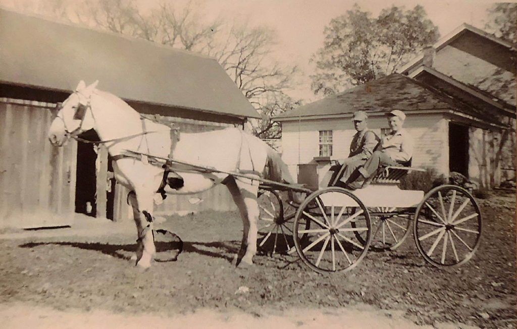 2 men in farm carriage with white hourse pose in front of barn and cream brick farmhouse with garage, ca. 1930s