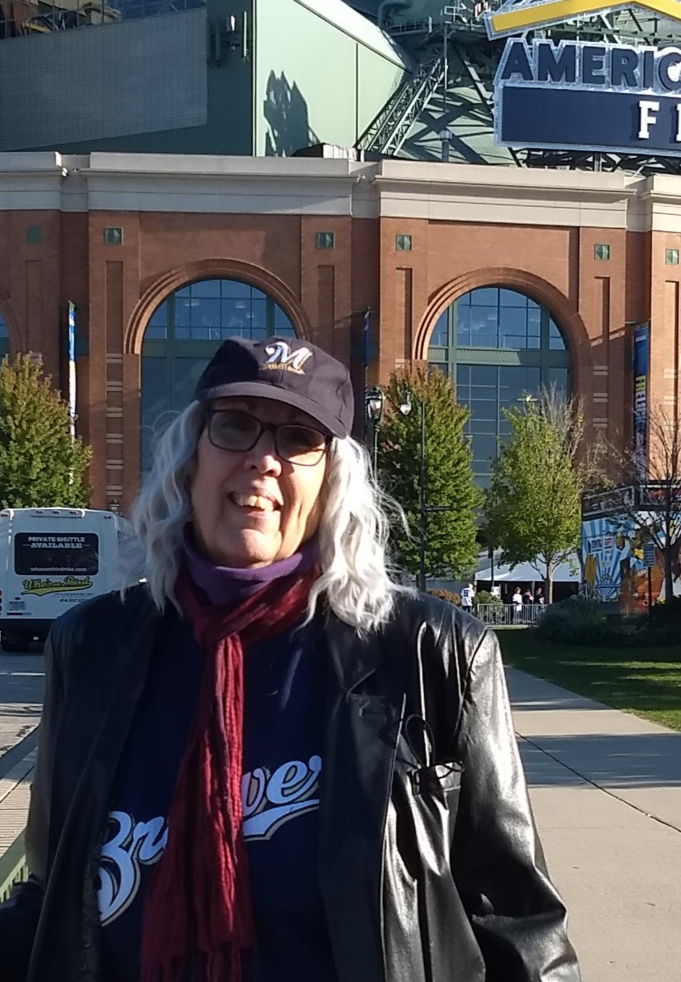 Tall female-presenting person with shoulder-length white wavy hair, wearing blue denim jacket, jeans, red scarf, and baseball cap, stands in front of American Family Field baseball stadium