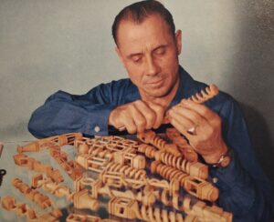 Middle-aged white man in blue denim shirt is carving a whimsy stick. Before him on a table are other whimsies he has carved.