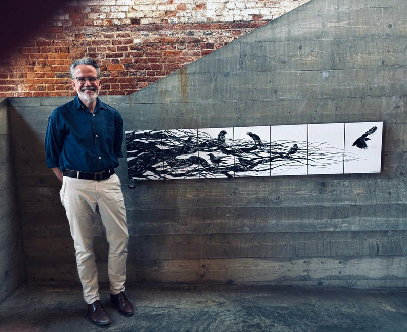 Stephen Richards, a tall, white middle-aged man wearing a navy button-down shirt and off-white trousers, stands next to his artwork titled